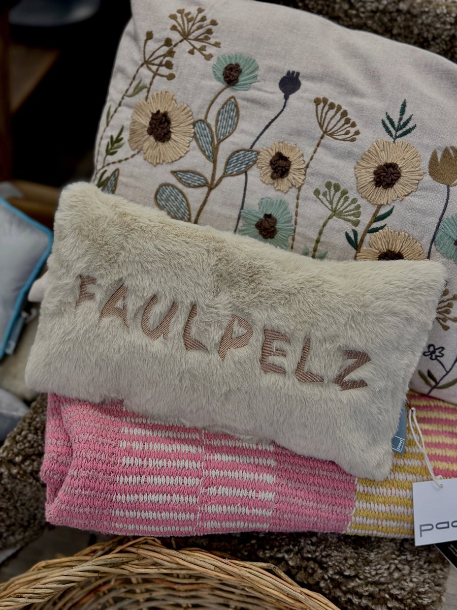 Kissenstapel in einem Korb: oben ein Kissen mit gesticktem Blumenmuster, davor ein flauschiges Kissen mit dem Schriftzug FAULPELZ, darunter Strickdecken in Rosa und Gelb