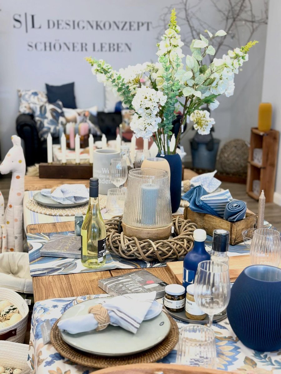 Gedeckter Tisch im Eingangsbereich mit Geschirr, Rattan-Untersetzern, Windlicht und weissen Blumen; im Hintergrund die Wand mit dem Schriftzug S|L Designkonzept - Schoener Leben